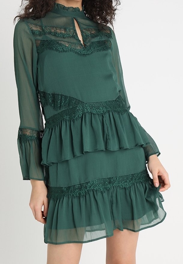 Mörkgrön klänning med långa ärmar, lager av volanger, spetsdetaljer och en nyckelhålsurklippning. Halvgenomskinligt tyg med texturerade detaljer.