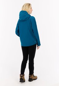 Mėlynas vandeniui atsparus džemperis su kapišonu, su laisvu siluetu ir ilgomis rankovėmis. Dėvimas su juodomis kelnėmis ir rudais batais, akcentuojantis lauko funkcionalumą.