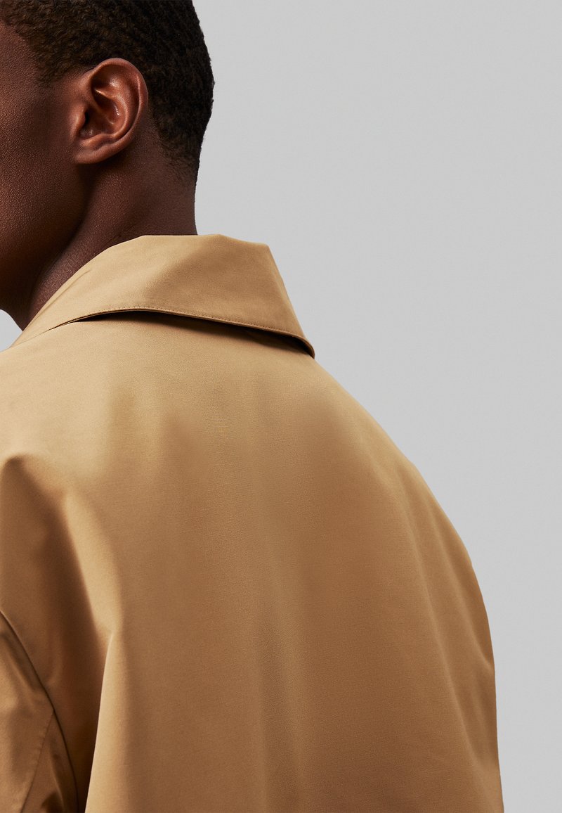 Brązowy, gładki płaszcz z dużym kołnierzem, pokazany z tyłu. Cechy obejmują czystą sylwetkę i minimalistyczny design.