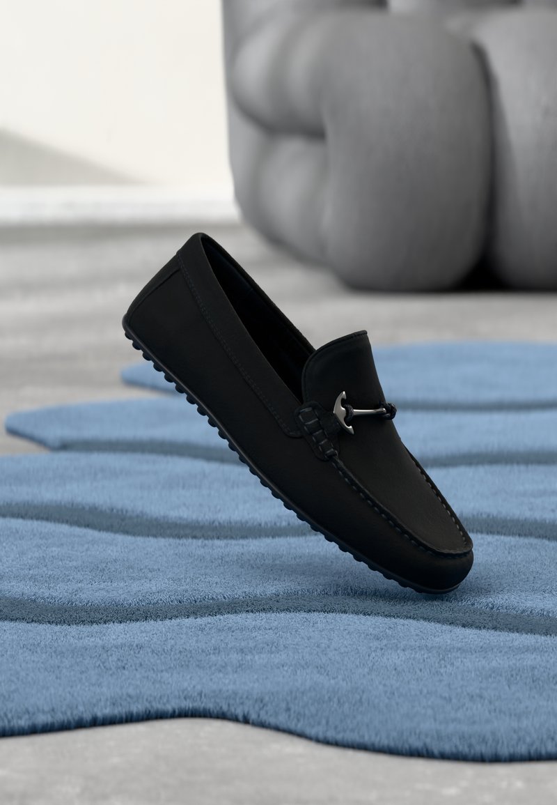 Mocassin en cuir noir avec un détail métallique argenté, flottant au-dessus d'un tapis bleu texturé, avec un fauteuil gris flou en arrière-plan.
