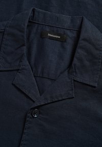 Chemise en lin bleu foncé avec un col cranté, un bouton visible et une étiquette noire avec un texte blanc "Matinique" à l’intérieur du col.