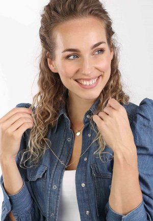 Gouden hartpendant ketting gedragen over een denim shirt. De ketting heeft een delicate schakel met een kleine ronde accen bij de hanger.
