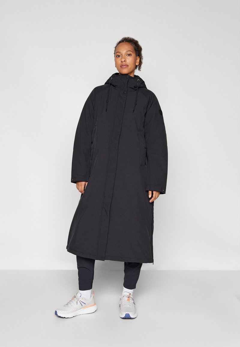 Lång svart parka med huva, dragkedjeslutning och sidofickor. Tillverkad av ett slätt material. Bärs över mörka byxor och ljusa sneakers.