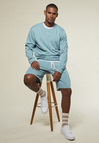 Mann, der auf einem Holzstuhl sitzt, trägt einen hellblauen Pullover und Shorts, weiße Sneakers und gestreifte Socken vor einem schlichten Hintergrund.