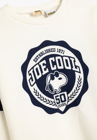 Beżowy dres z dużym granatowym okrągłym logo z napisem "JOE COOL", grafiką Snoopy i tekstem "50 LAT". Wykonany z bawełny.