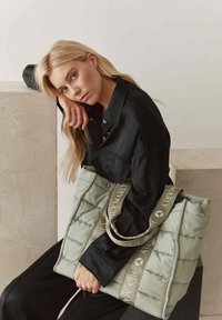 Gequilte groene tote bag met grijze handvatten, gebosseleerd met "HEYMARLY." Model in een zwart shirt poseert casual terwijl ze de tas vasthoudt tegen een lichte achtergrond.