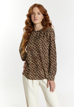 Blouse marron à manches longues avec un motif beige en boucle, dotée d'un col rond et de détails froncés aux poignets, associée à un pantalon crème.