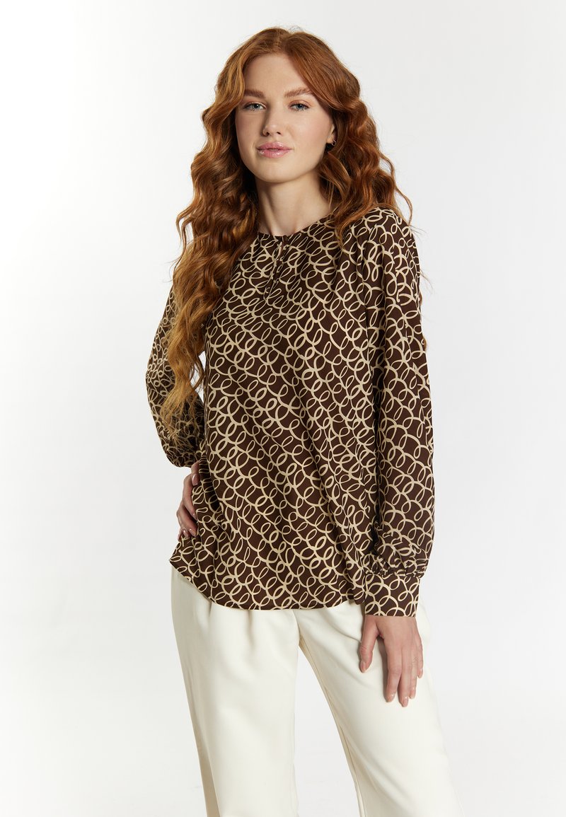 Brown long-sleeve blouse with a looping beige pattern, featuring a round neckline and gathered detail at the cuffs, paired with cream trousers.