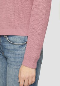 Roze gebreide trui met een textuurfinish. Geribbelde manchetten en zoom, gecombineerd met lichtblauwe denim. Focus op de mouwen en hand.
