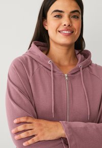 Lichtpaarse rits-hoodie met een drawstring capuchon, raglanmouwen en een zachte textuur, getoond met gekruiste armen tegen een neutrale achtergrond.