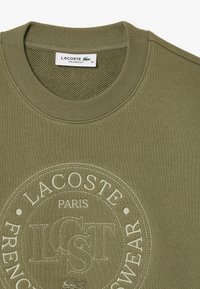 Sudadera verde oliva con cuello redondo y detalles de canalé. Presenta un logotipo circular y una marca bordada en la parte frontal. Ajuste relajado.