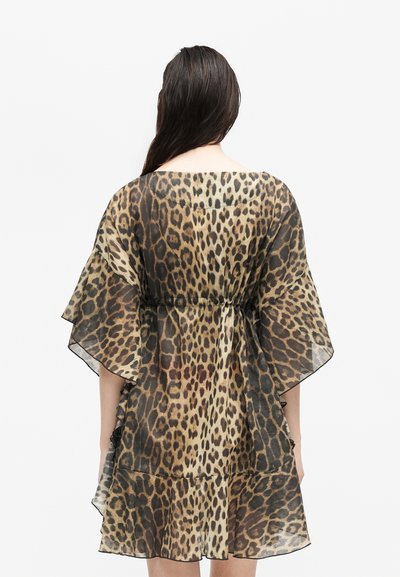 Γυναίκα με μακριά σκούρα μαλλιά, που φοράει ένα διαφανές, φαρδύ φόρεμα με λεοπάρ μοτίβο και φαρδιά μανίκια, φωτογραφημένη από πίσω με λευκό φόντο.