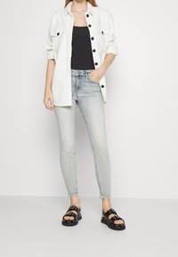 Camisa blanca de botones, top negro, jeans ajustados de mezclilla azul claro, sandalias negras con remaches. Capa casual con colores y texturas contrastantes.