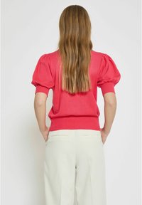 Růžový pletený top s balónovými rukávy a žebrovaným lemem, spárovaný s krémovými kalhotami s zadními kapsami. Tkanina vypadá měkke a jemně.