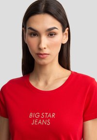 Raudona medvilninė marškinė su apvaliu iškirpimu ir trumpomis rankovėmis. Priekyje išsiuvinėtas užrašas "BIG STAR JEANS" auksinės spalvos. Lygi tekstūra.