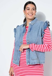 Spijkervest met gerimpelde schouders, lichtblauwe kleur, voorkanten met zakken en zilveren knopen, gedragen over een rood-wit gestreepte jurk met lange mouwen.