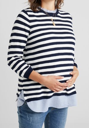 Vrouw in een blauw-wit gestreepte trui en een blauwe spijkerbroek, zachtjes haar zwangere buik met beide handen ondersteunend.