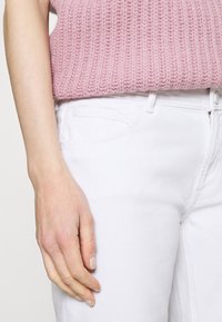 Jean en denim blanc à texture lisse, présentant un design standard à cinq poches. Au-dessus, un pull en tricot rose clair avec un motif côtelé.