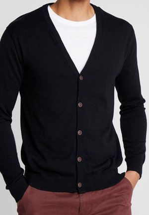 Mann trägt einen schwarzen, geknöpften Cardigan über einem weißen T-Shirt mit den Händen in den Taschen dunkelroter Hose.