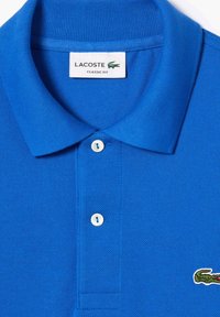 Modrá bavlnená polokošeľa s rebrovaným golierom, trojbuttonovou légou a vyšitým zeleným logom krokodíla na ľavej hrudi. Klasický strih.