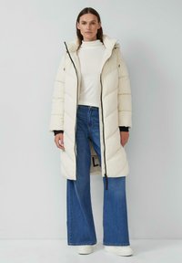 Krem krāsas pildīts garais mētelis ar horizontālām quiltēšanām, ar rāvējslēdzēju priekšā un regulējamu kapuci. Apvienots ar platām biksēm zila krāsā un baltām kedām.