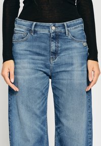 Blaue Denim-Jeans mit lockerer Passform, zwei Fronttaschen, Knopfverschluss und dezenten Auswaschungen. Etikett auf der Rückseite sichtbar.