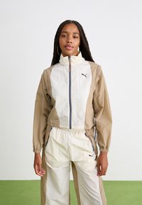 Lett jakke med beige og kremfargede paneler, hel glidelås, stående krage, elastiske mansjetter og logodetaljer. Kombinert med matchende kremfargede bukser.