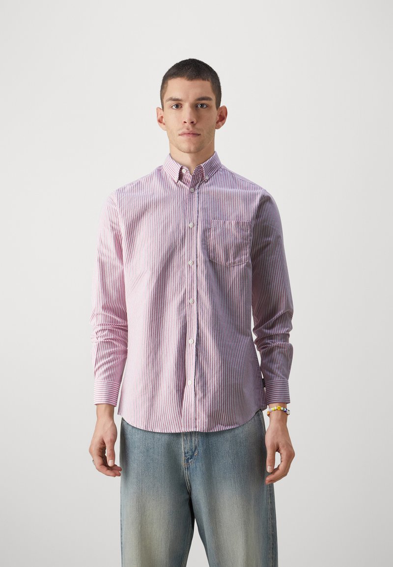 Camisa a rayas en rojo y blanco, con un bolsillo en el pecho y mangas largas. Combinada con jeans de mezclilla de un azul claro, ligeramente desgastados.