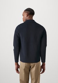 Navy knit pullover with a collar and long sleeves. Smooth texture, simple design. Worn with light-colored trousers.