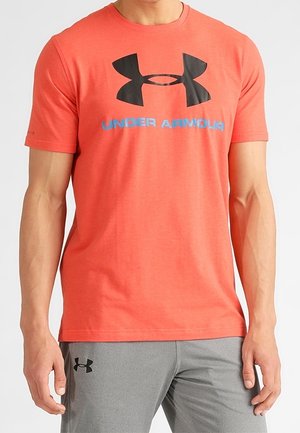 Mężczyzna noszący koralowy T-shirt Under Armour z czarnym logo i napisem, w połączeniu z szarymi szortami z małym czarnym logo Under Armour.