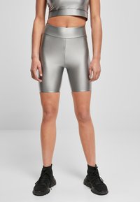 Kobieta ubrana w błyszczące, srebrne szorty rowerowe z wysokim stanem oraz dopasowany top, w połączeniu z czarnymi sportowymi butami, stojąca na szarym podłożu.