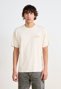 Beige puuvillane T-särk lühikeste varrukatega; vasakul rinnal on kerge pruun "Dickies" logo. Kandmiseks sobivad hallid teksad.