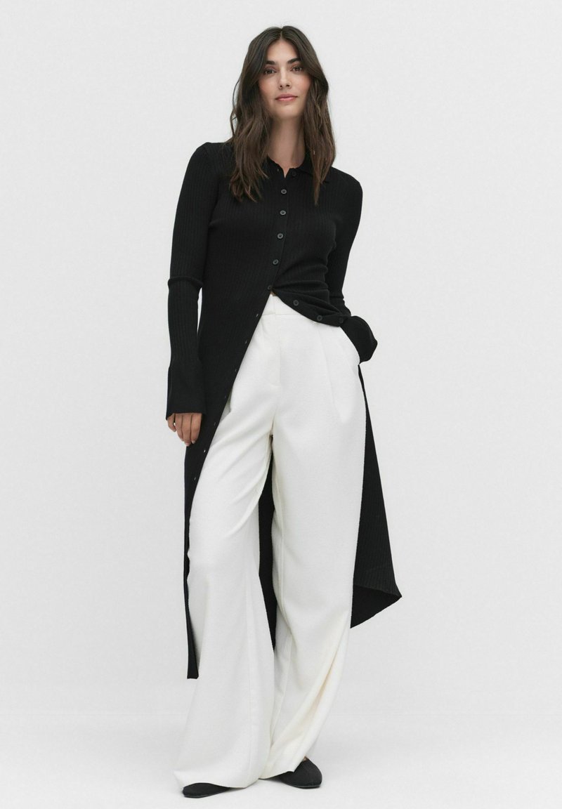 Cardigan noir côtelé avec fermeture à boutons, manches longues et côtés fendus, associé à un pantalon blanc à jambes larges en tissu doux.
