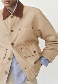 Beige canvas jacket with a brown corduroy collar, four front pockets, and snap buttons. Worn over a light blue striped shirt.
