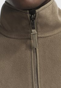 Fleece-Jacke mit Reißverschluss in Olivgrün, ausgestattet mit einem hohen Kragen, strukturiertem Material und einem kontrastierenden Reißverschluss mit dem Markennamen.