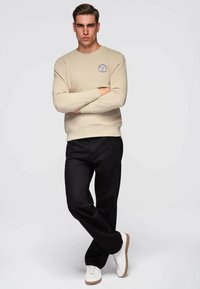 Beige sweatshirt met een ronde logo-badge, geribbelde manchetten en zoom; gecombineerd met zwarte broek en witte sneakers. Simpele, casual outfit.