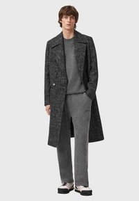 Szary, teksturowany płaszcz nałożony na dopasowany sweter i spodnie, do tego kontrastujące czarno-białe trampki oraz subtelne logo na spodniach.