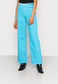 Calças de veludo cotelê azul-claro, com cintura alta, textura riscada verticalmente e bolsos frontais, combinadas com botas pretas de salto.
