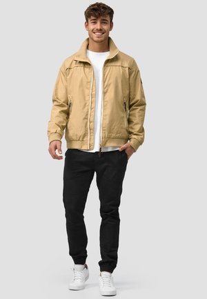 Młody mężczyzna uśmiecha się, ubrany w beżową kurtkę na białą koszulę, czarne spodnie i białe trampki, stoi na jasnoszarym tle.