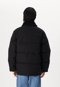 Schwarze Daunenjacke mit einem fleecegefütterten Kragen, übergroßer Schnitt und gestepptem Design. Trägt sie über hellblauen Jeans. Rückansicht gezeigt.