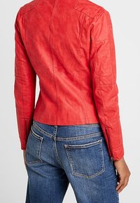 Giacca di pelle rossa con dettagli cuciti su spalle e maniche; design aderente con polsini con zip; indossata con jeans di denim blu.