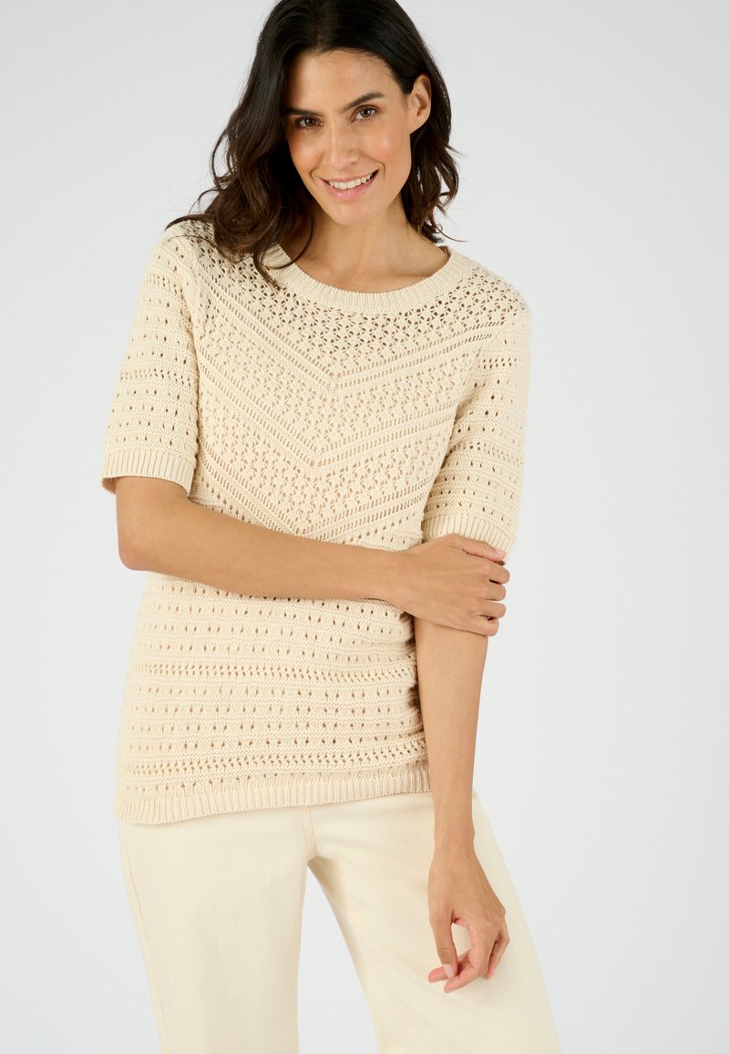 Pull à manches courtes couleur crème avec un design en maille ouverte texturée, doté d'un col rond et d'une finition côtelée. Porté avec un pantalon clair.