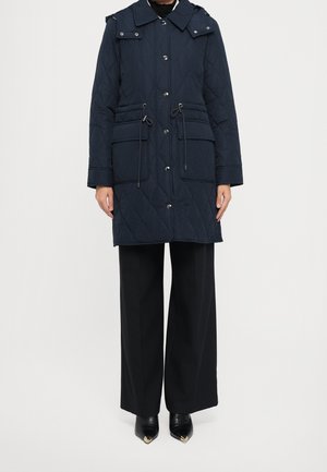 Femme portant un manteau matelassé bleu marine avec poches avant et cordons de serrage, un pantalon noir à jambes larges et des chaussures noires à bouts pointus ornées d'embouts dorés.