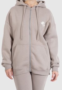 Felpa zip beige chiaro in morbido tessuto con tasche frontali, cappuccio con cordino e un piccolo logo bianco sul petto sinistro.