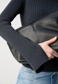 Borsa a mano nera in pelle tenuta tra le mani, caratterizzata da una texture liscia, bordi morbidi e cuciture sottili. Indossata con un top blu navy a coste.