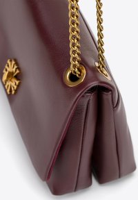 Bordová kožená kabelka s chevronovým vzorem, obsahující zlatý řetízek a ozdobné kování na přední straně. Texturovaný povrch.