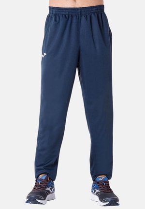 Mann trägt dunkelblaue Sporthose und schwarze, blaue und orange Laufschuhe und steht vor einem schlichten Hintergrund.