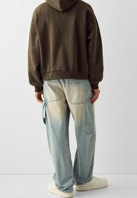 Felpa con cappuccio marrone, jeans cargo celeste sbiadito con tasche applicate e sneakers bianche. Tessuto morbido, vestibilità rilassata e stile casual.