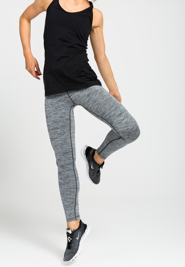 Personne portant un débardeur noir, des leggings gris et des baskets noires Nike, levant une jambe pliée au genou sur fond blanc.