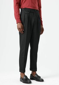 Schwarze Nadelstreifenhose mit maßgeschneiderter Passform, hoher Taille, zwei Knöpfen am Bund und umgeschlagenen Säumen. Kombiniert mit schwarzen Loafers.